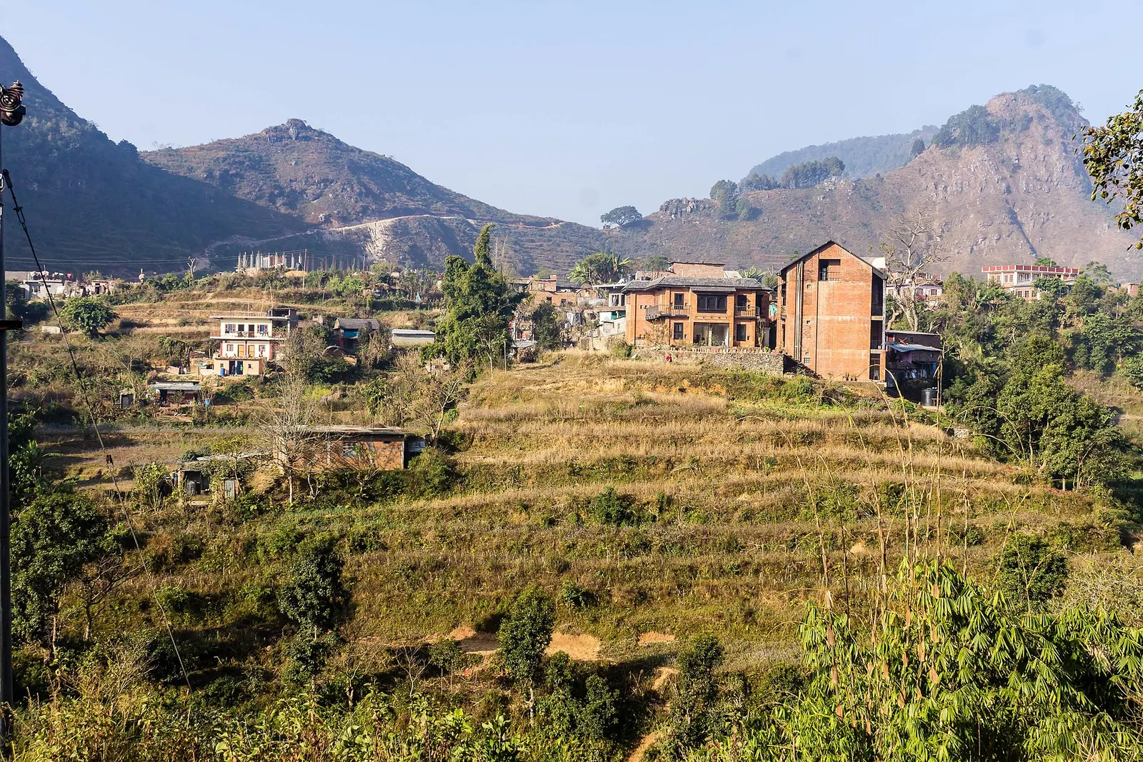 Terraced hillside fields below brick houses in Bandipur, with rugged hills behind