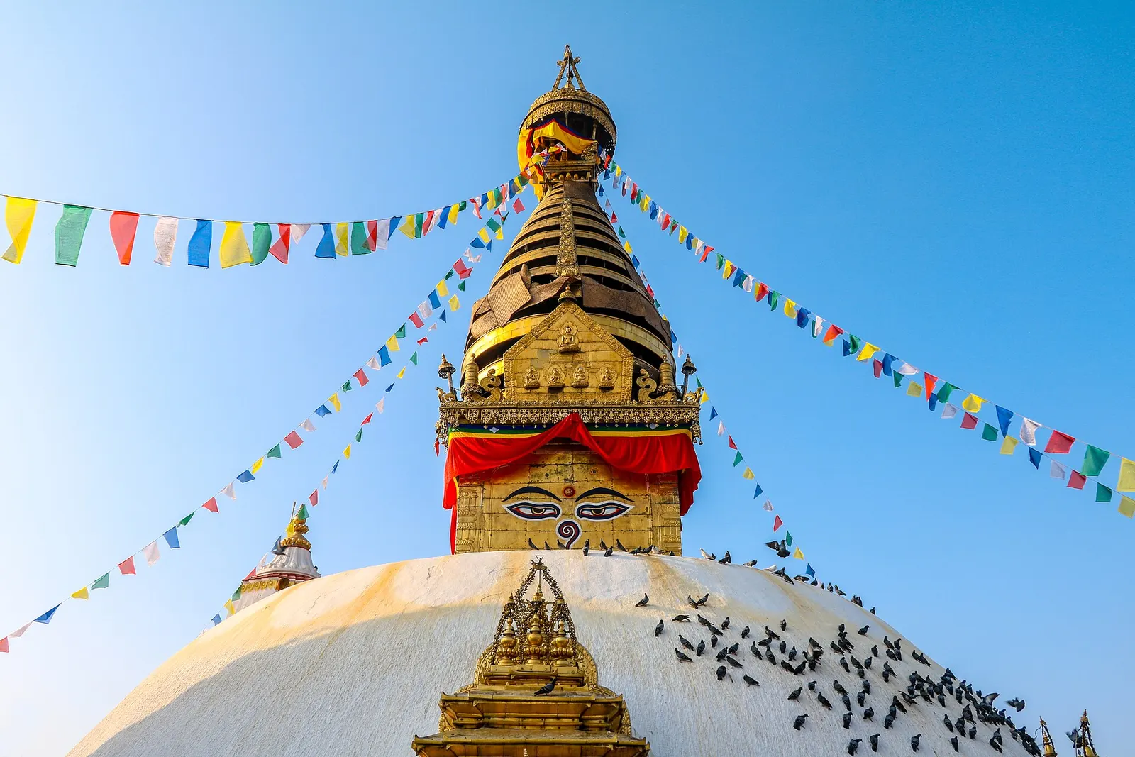 Golden spire of Swayambhunath Stupa with painted Buddha eyes and strings of prayer flags against a clear blue sky