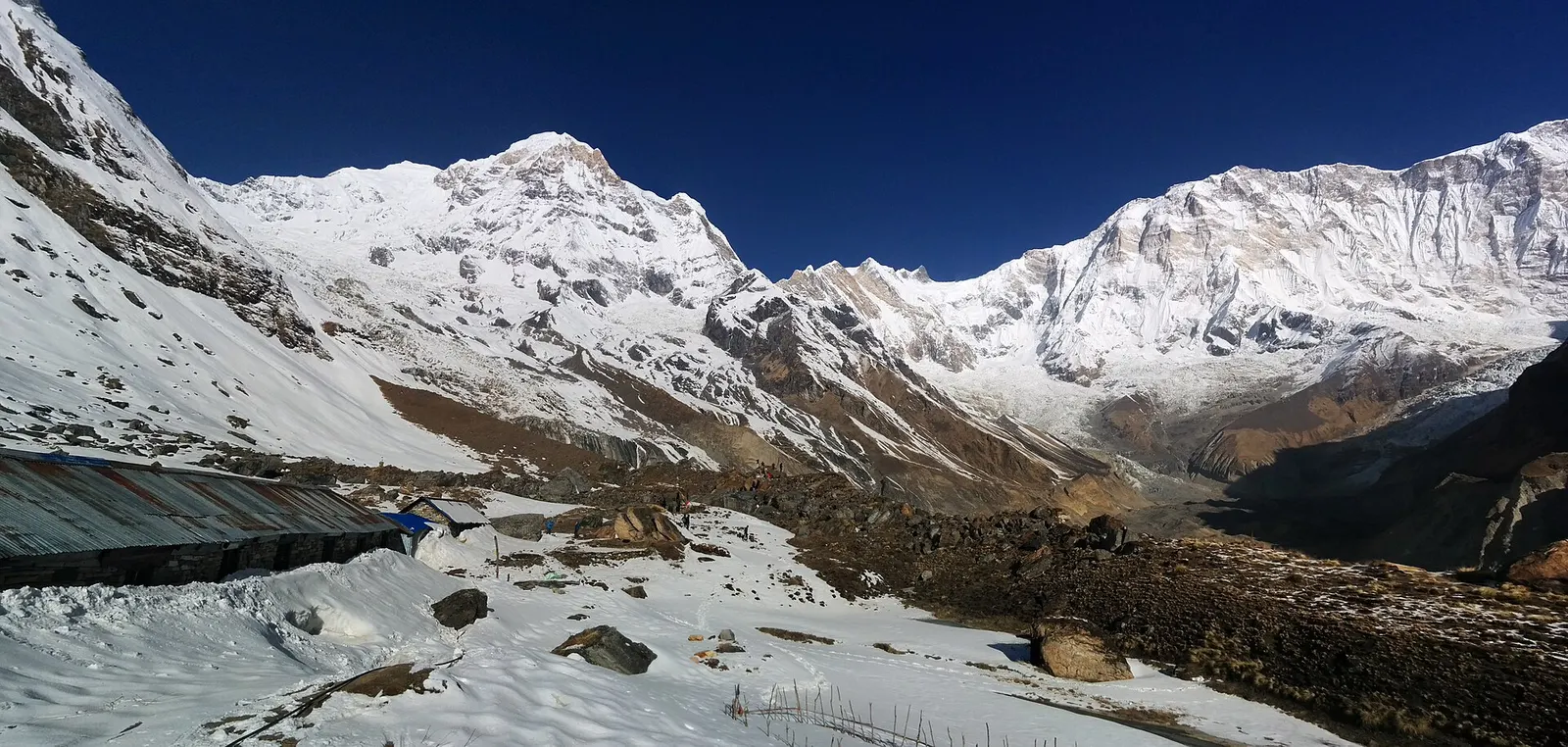 Snow-covered ground and lodge roofs at Annapurna Base Camp below Annapurna I and its neighbouring peaks under a deep blue sky.