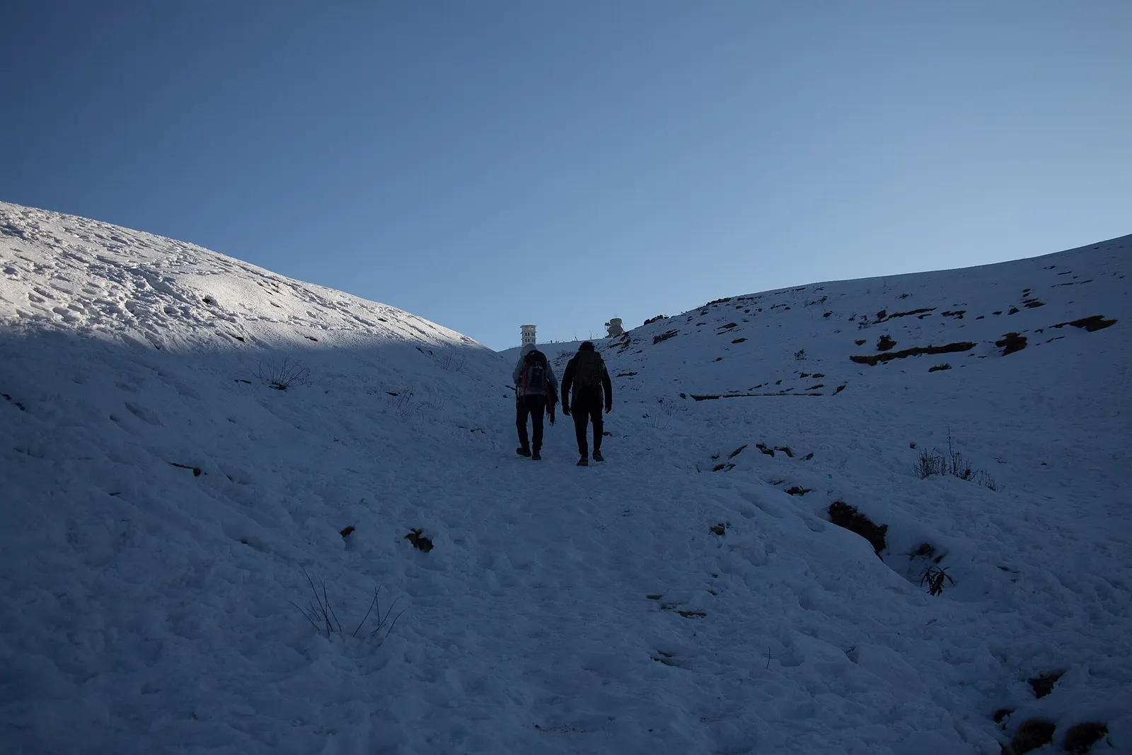 Two hikers walking up a snow-covered grassy slope towards the Sailung hilltop.