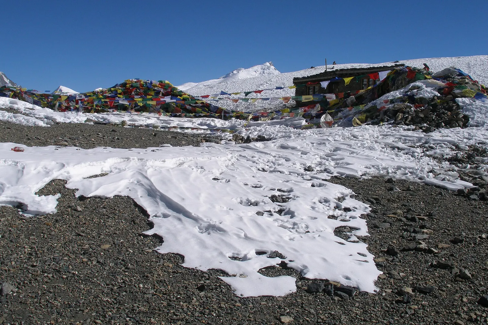 Prayer flags and a small stone hut at the Thorong La, with patches of snow on grey scree under a blue sky.