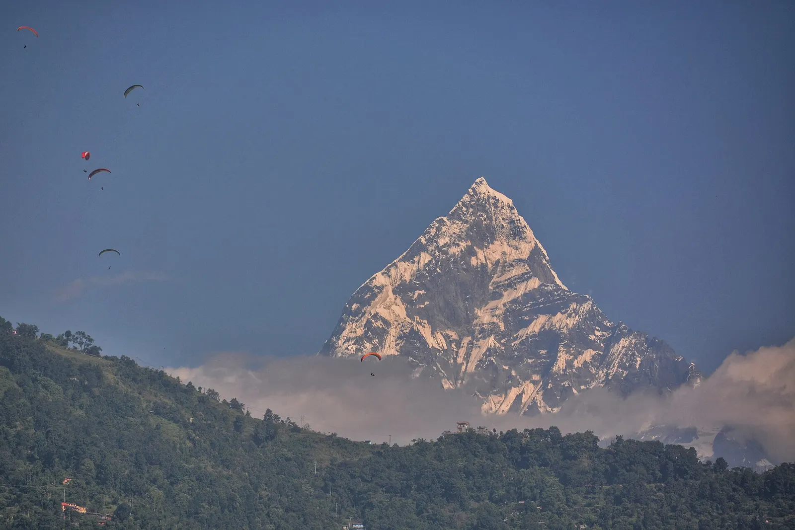 The pointed summit of Machhapuchhre glowing above a band of cloud and forested hills, with paragliders in the sky over Pokhara