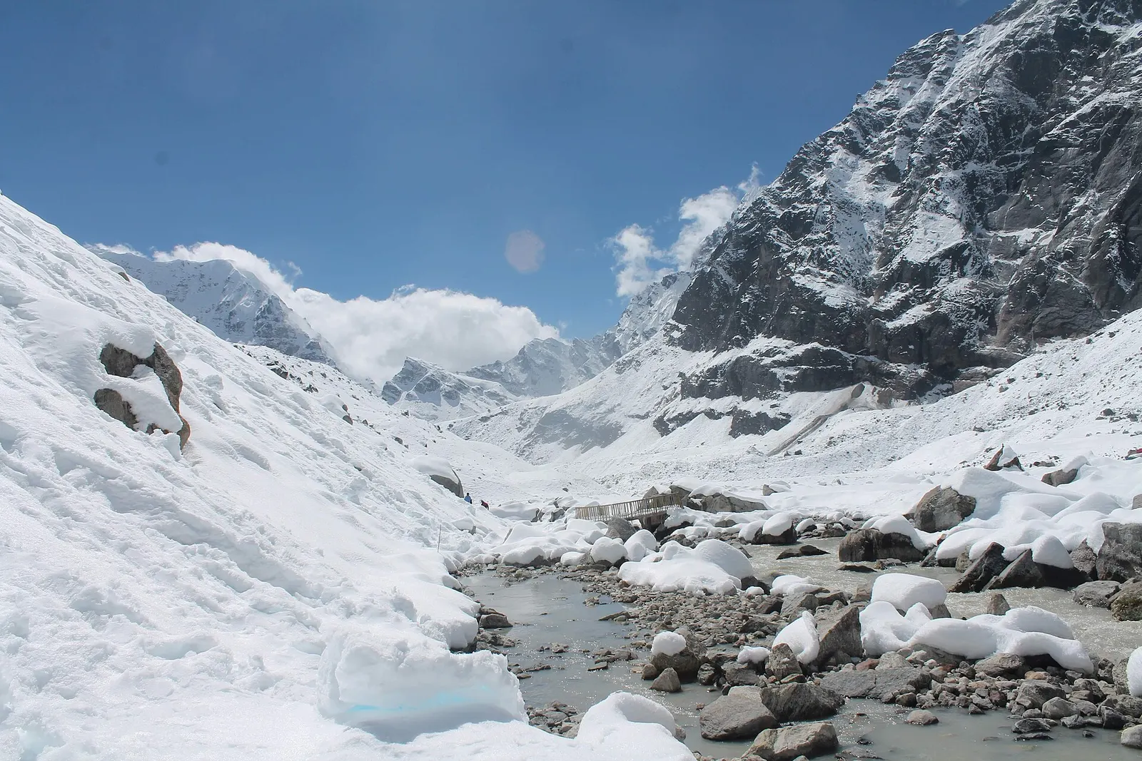 A snow-covered valley with a glacial stream and small footbridge at Makalu Base Camp, below steep rock walls.