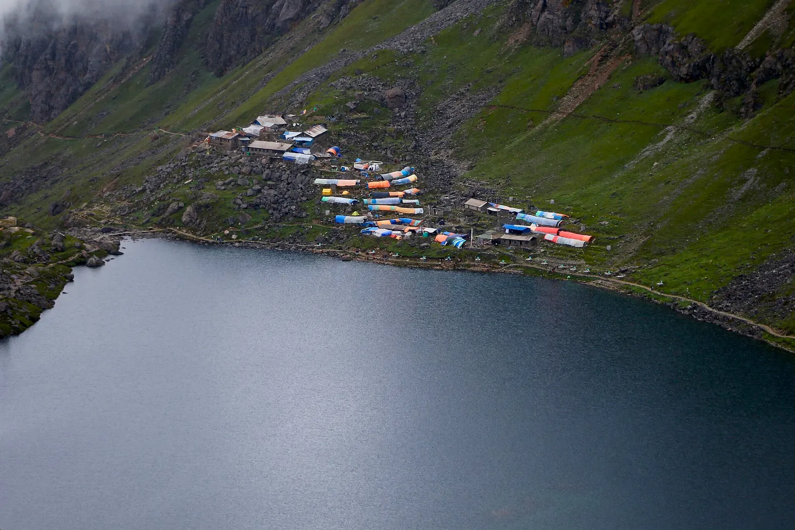 The dark water of Gosaikunda lake with a cluster of lodges with coloured roofs on the green slope above its shore.